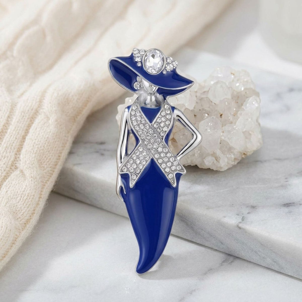 A fashion-themed enamel pin shaped like a woman in a blue hat and dress, decorated with silver-tone rhinestone-embellished details, displayed on a white marble slab with a cream knit fabric and crystal cluster in the background.