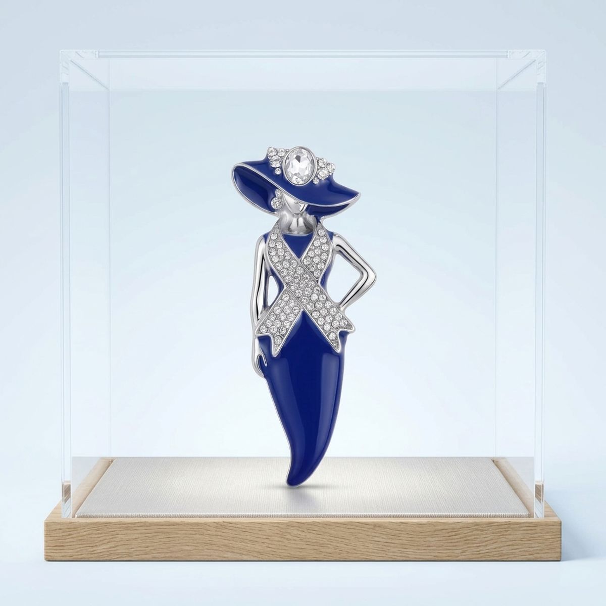 A fashion jewelry piece of a woman in a blue dress and hat with silver crystal-embellished details, displayed inside a clear acrylic case on a light wood base.