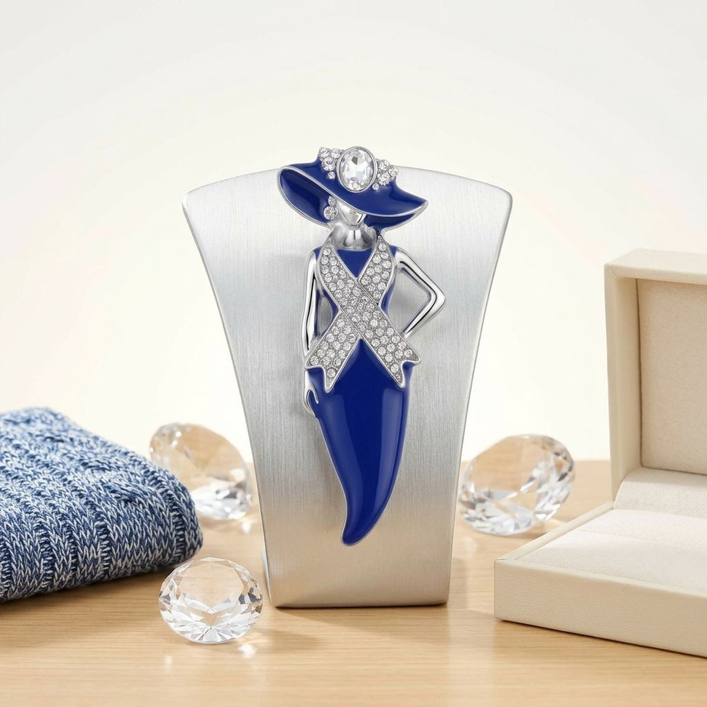 A decorative fashion figurine of a woman in a blue hat and blue dress with silver crystal-embellished details, displayed on a silver stand on a wooden surface with a blue knit fabric, clear faceted stones, and an open jewelry box nearby.