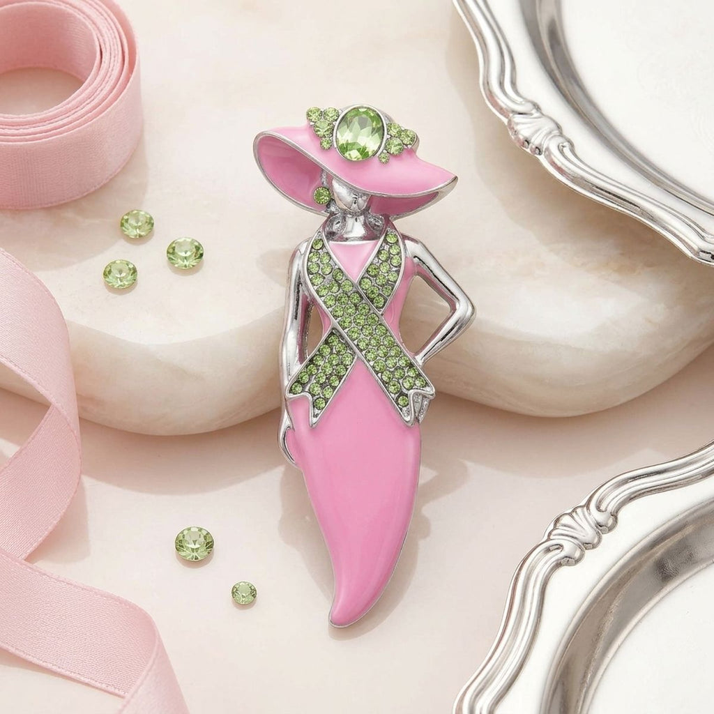 A pink and silver awareness ribbon brooch shaped like a woman in a hat, embellished with light green gemstones, displayed on a light pink marble surface with pink ribbon and silver trays.