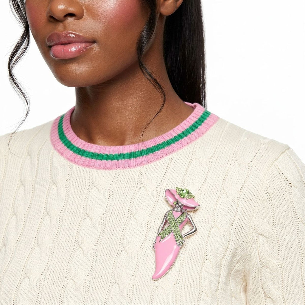 A close-up of a model wearing a cream cable-knit sweater with pink and green striped trim, featuring a pink and green high-heel brooch pinned to the chest.