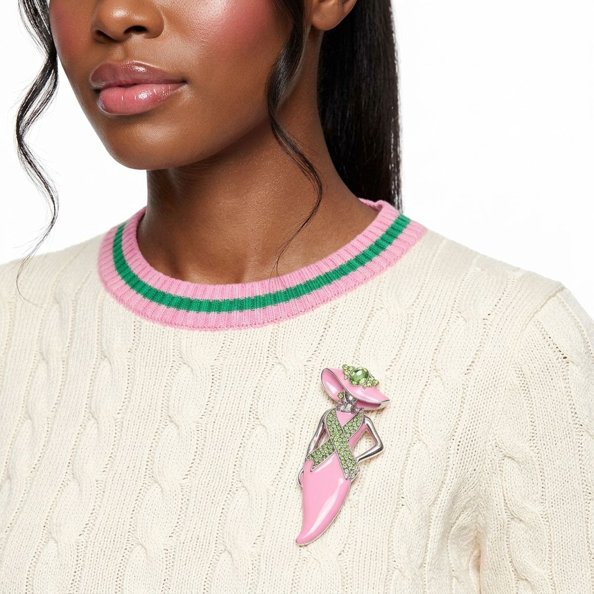 A close-up of a model wearing a cream cable-knit sweater with pink and green striped trim, featuring a pink and green high-heel brooch pinned to the chest.