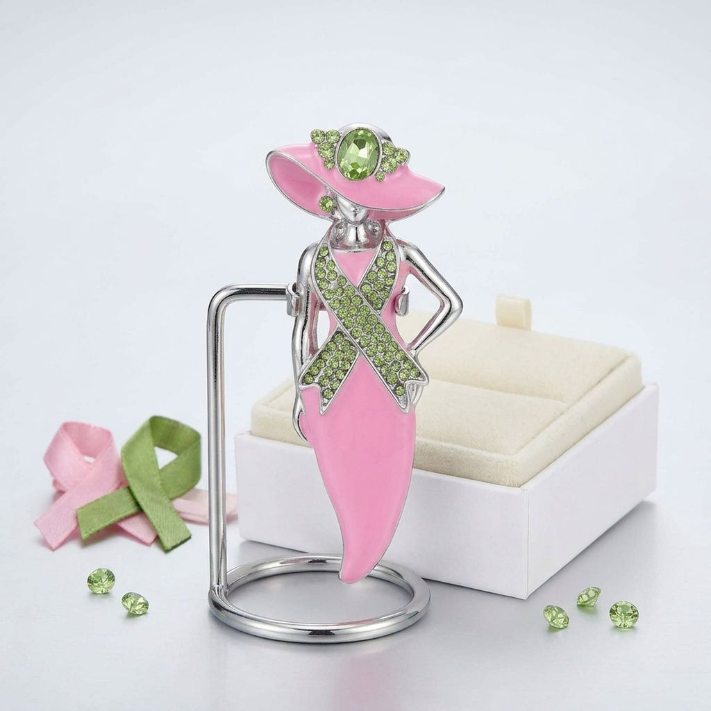 A pink and silver awareness ribbon brooch shaped like a woman in a hat, displayed on a stand with a cream jewelry box, pink and green ribbon props, and small green rhinestones on a white background.