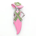 A pink and silver awareness ribbon brooch shaped like a woman wearing a pink hat, decorated with green rhinestones and a green gemstone, on a white background.