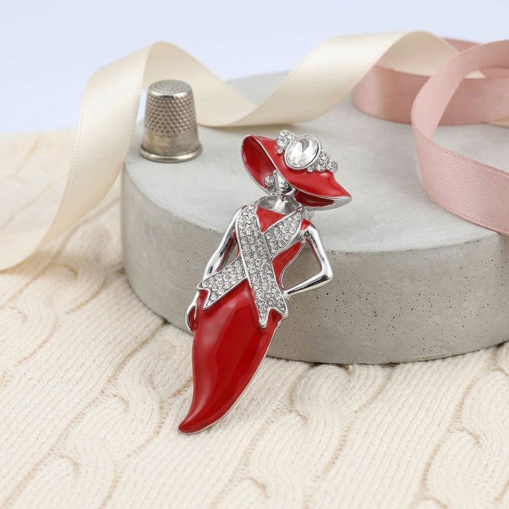 A red and silver fashion dress-shaped brooch with rhinestone accents, displayed on a gray stone pedestal with cream and pink ribbons.