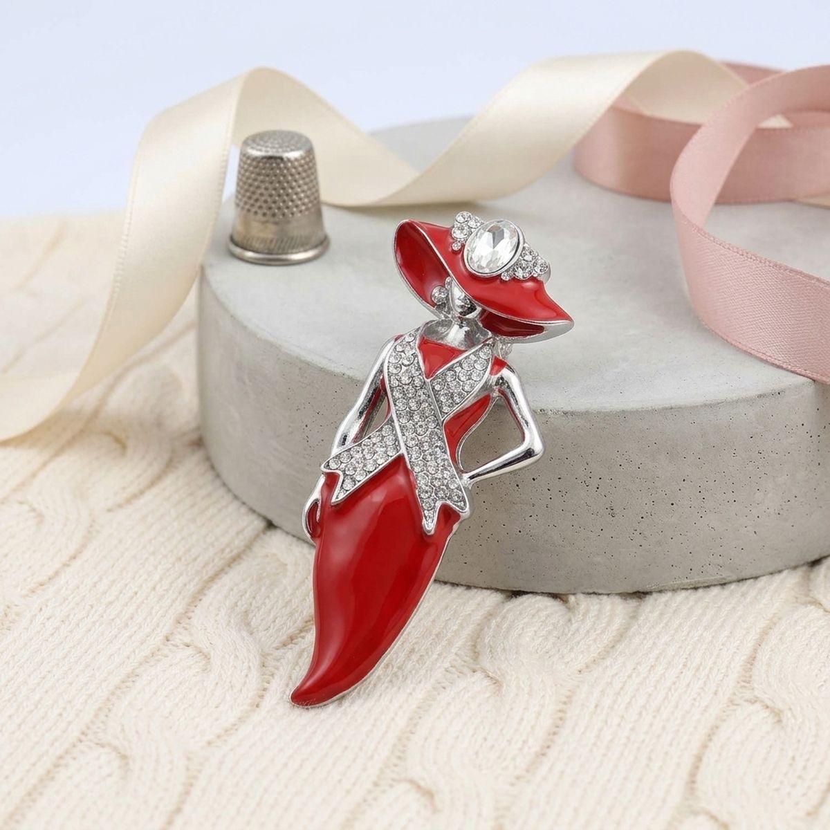 A red and silver fashion dress-shaped brooch with rhinestone accents, displayed on a gray stone pedestal with cream and pink ribbons.