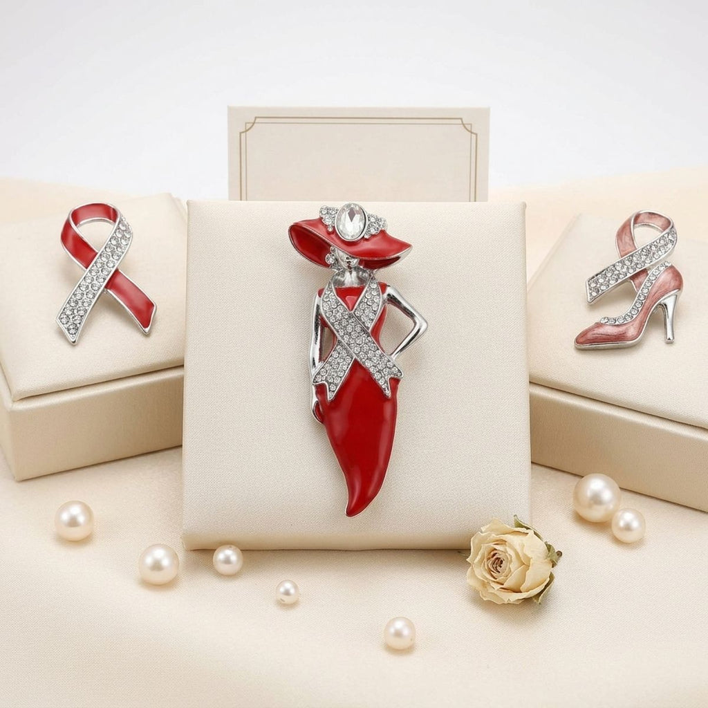 A jewelry display featuring a central red and silver dress-shaped brooch on a cream stand, with matching ribbon and high-heel jewelry pieces arranged on boxes around it. Pearls and a small rose are placed in front on a light beige surface.