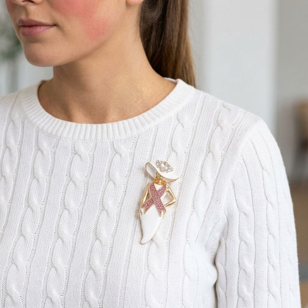 White enamel hat lady brooch in gold tone with pink rhinestone breast cancer awareness ribbon and crystal hat accent, pinned on a woman's white cable-knit sweater