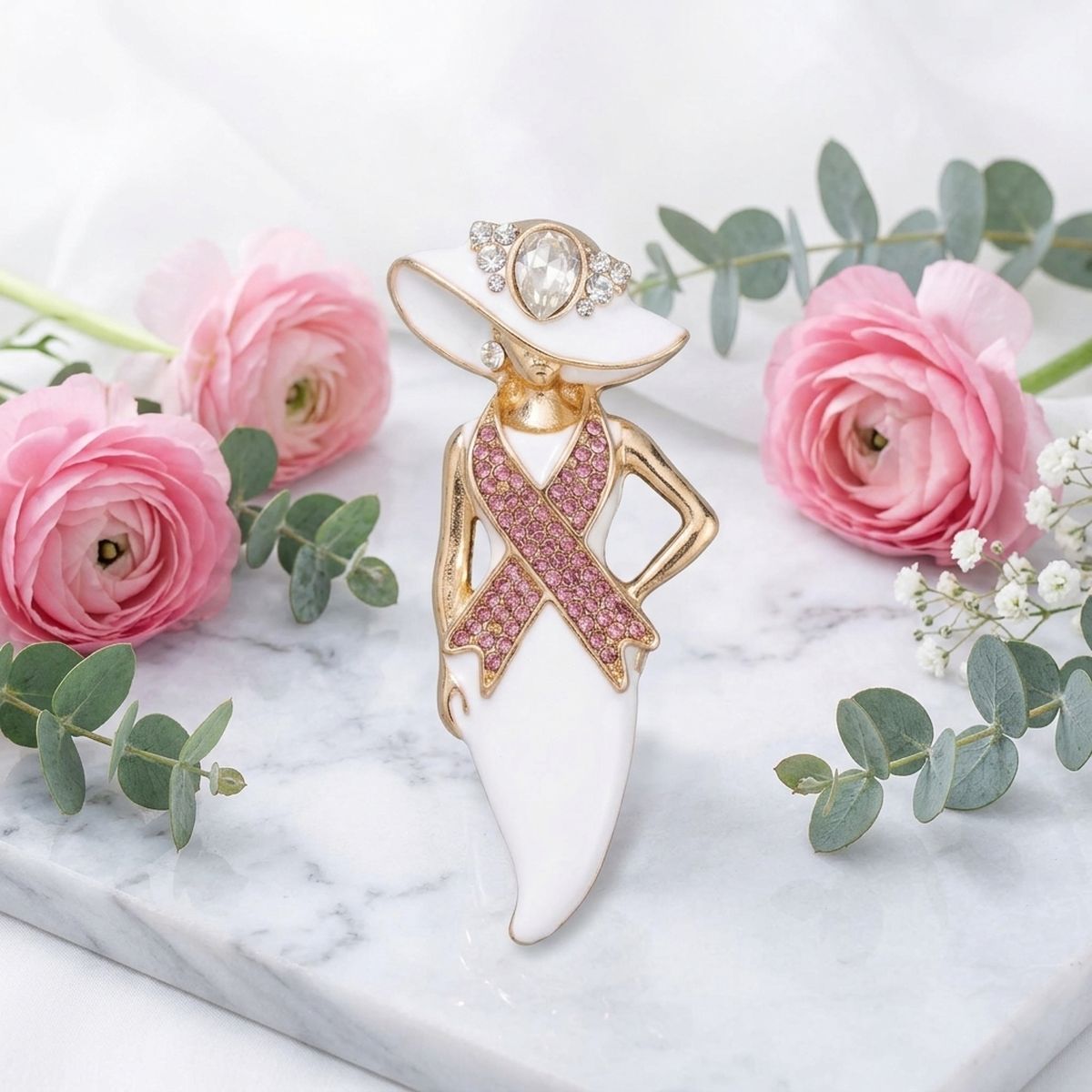 White and gold breast cancer awareness brooch displayed on white marble with pink ranunculus flowers and eucalyptus