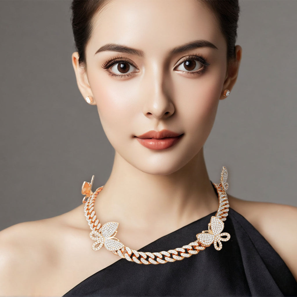 Woman wearing gold rhinestone butterfly Cuban chain necklace with pave butterfly charms and small stud earrings, black one-shoulder top, gray background