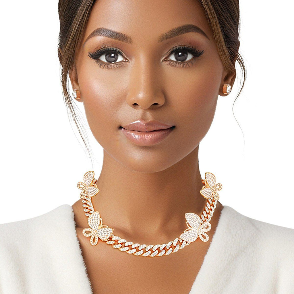 Woman wearing gold rhinestone-iced butterfly Cuban chain necklace with pave butterfly stations and small square stud earrings, white blazer, white background