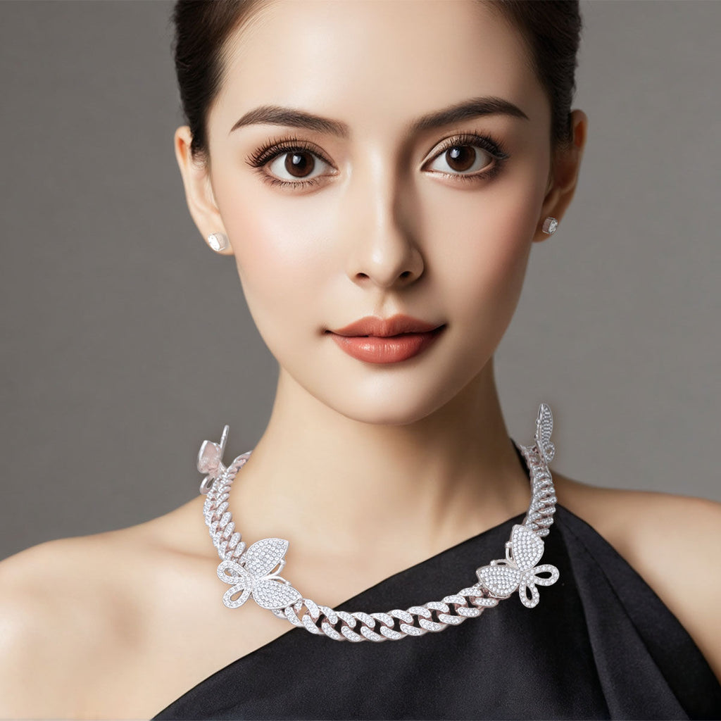 Woman wearing rhinestone-iced silver butterfly Cuban chain necklace with pave butterfly stations and small stud earrings, black one-shoulder top, gray background