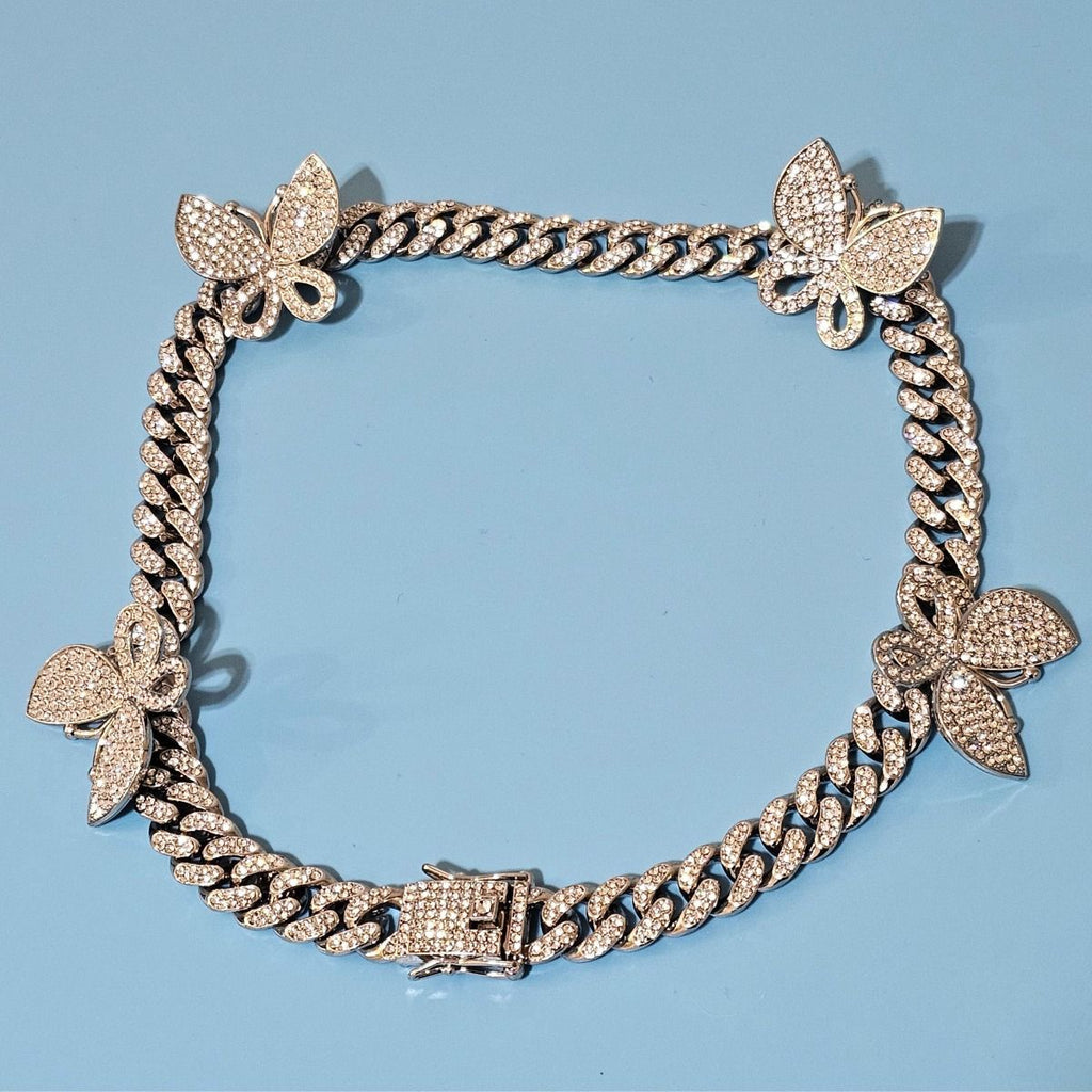Close-up flat lay of silver rhinestone-iced butterfly Cuban chain necklace with five pave butterfly stations and push-lock box clasp, light blue background