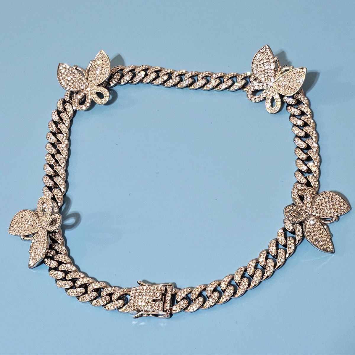 Close-up flat lay of silver rhinestone-iced butterfly Cuban chain necklace with five pave butterfly stations and push-lock box clasp, light blue background