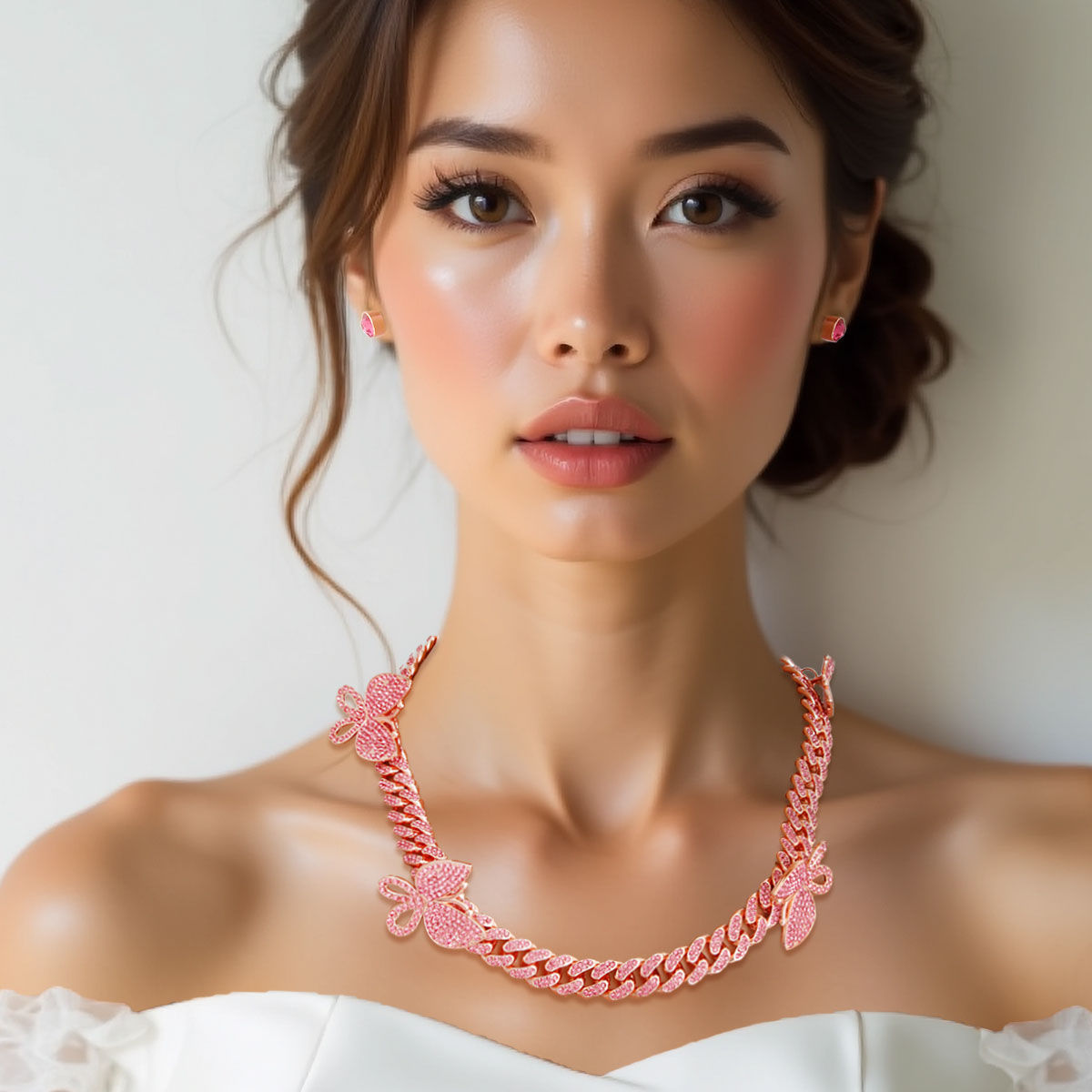 Woman wearing pink rhinestone-iced rose gold butterfly Cuban chain necklace with five pave butterfly stations and matching pink butterfly stud earrings, white off-shoulder top, beige background