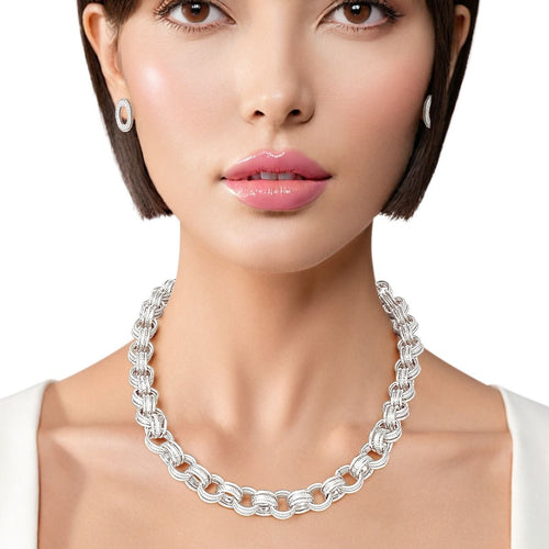 A woman with light skin, short dark bob haircut, and pink glossy lips poses against a white background wearing a white sleeveless top. She wears a chunky silver rhodium tone triple ring rolo chain choker necklace sitting at the collarbone, with matching small open circle ring stud earrings with ribbed texture detailing visible on both ears.