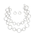 Full set laid out on white background — double layer chunky silver ring link necklace with mixed small and large open circular rings connected by rectangular hammered barrel links, lobster clasp and extender chain visible, with two matching small circular fish hook earrings centered between the necklace layers