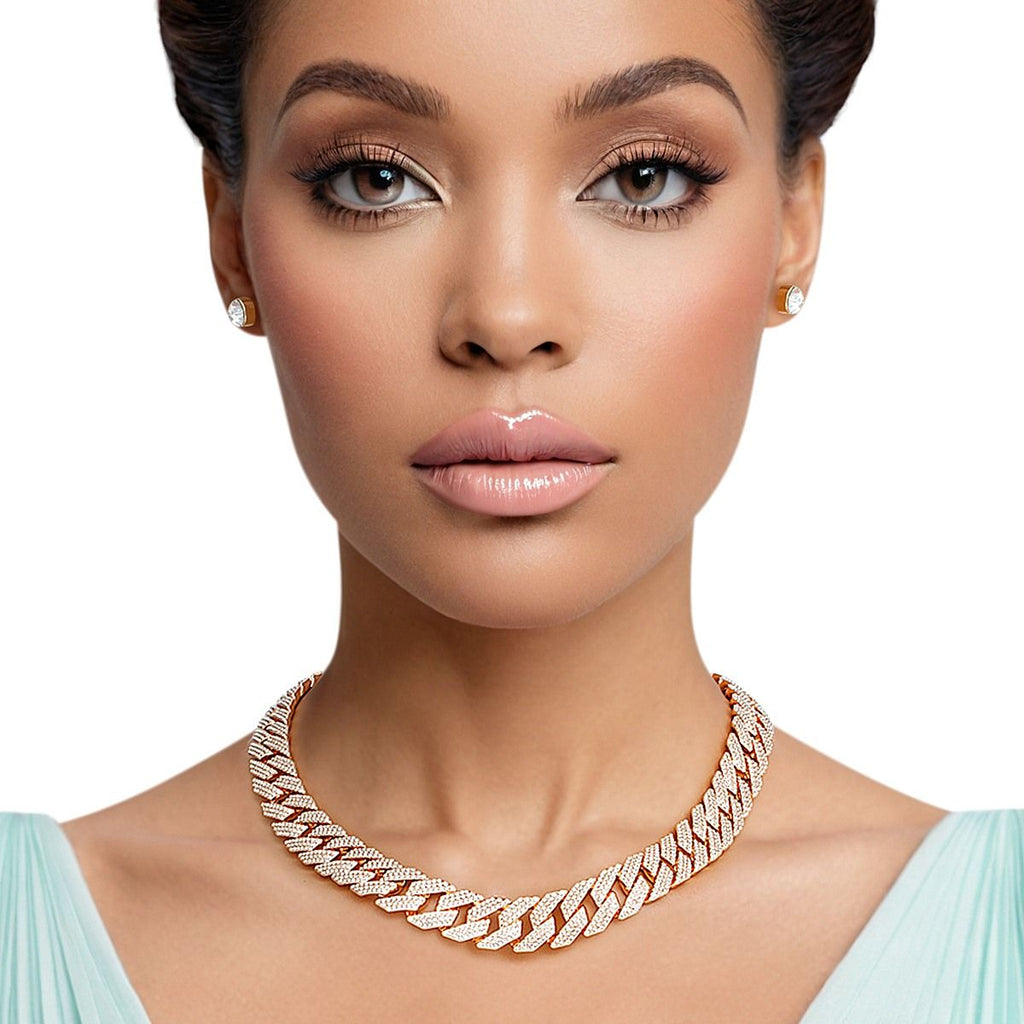 A woman with medium-dark skin, dark upswept hair, and bold eye makeup poses against a white background wearing a wide 14mm fully pave clear rhinestone Miami Cuban chain link necklace in gold tone at the collarbone, paired with matching small round clear rhinestone stud earrings with gold tone settings.