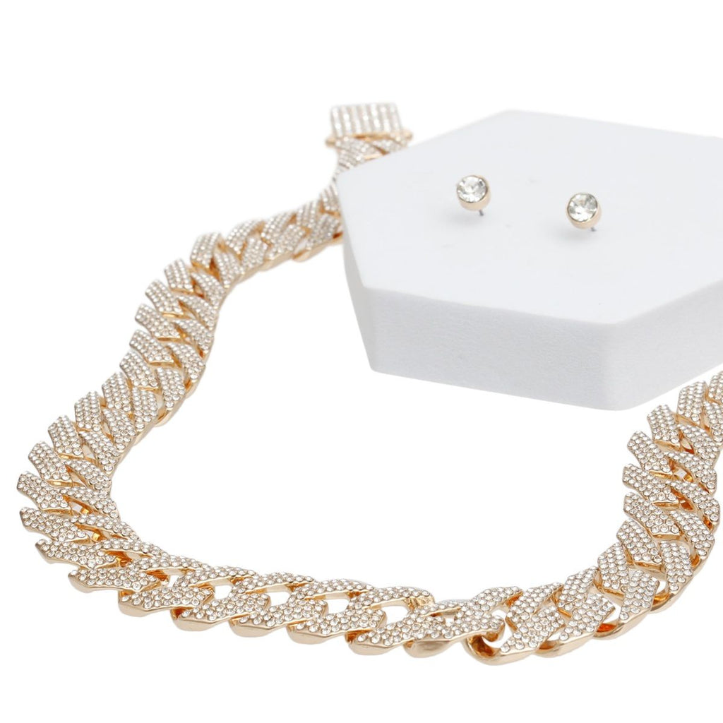 Angled close-up of a wide 14mm fully pave clear rhinestone Miami Cuban chain link necklace in gold tone draped over a white hexagonal display stand. Two small round clear rhinestone stud earrings with gold tone butterfly post backings are displayed on top of the white stand beside the necklace clasp.