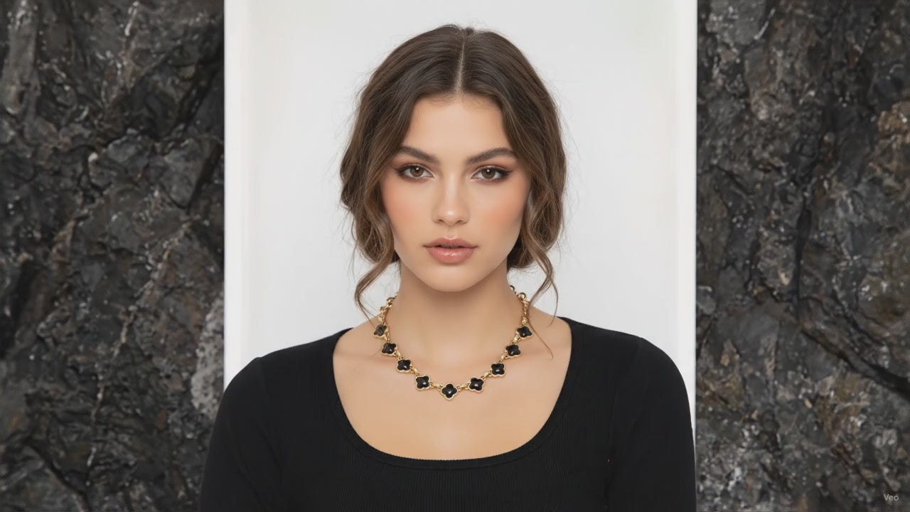 Black quatrefoil stone bold link chain necklace in worn gold-tone with repeating four-leaf motifs and lobster clasp extender, flat lay on white background