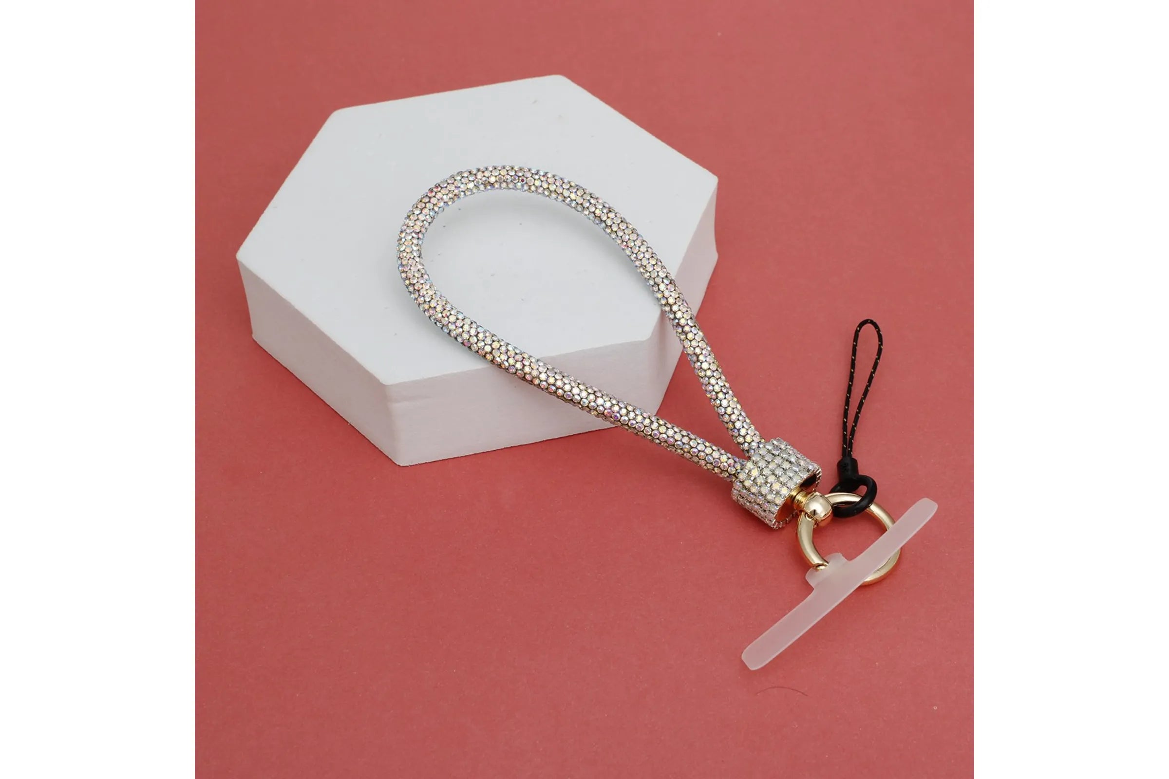 Aurore borealis iridescent rhinestone-encrusted rope phone wrist strap draped over white hexagonal display prop on coral red background, showing full loop, clear rhinestone square barrel wrap, round gold carabiner clasp, white plastic card insert, and black cord loop