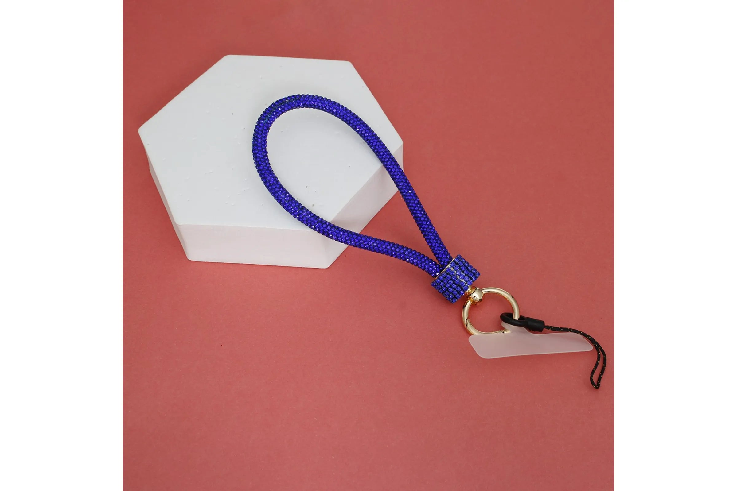 Royal blue rhinestone rope phone wrist strap draped over white hexagonal display prop on red background, showing full loop, blue rhinestone barrel knot wrap, gold carabiner clasp, clear plastic card insert, and black cord loop