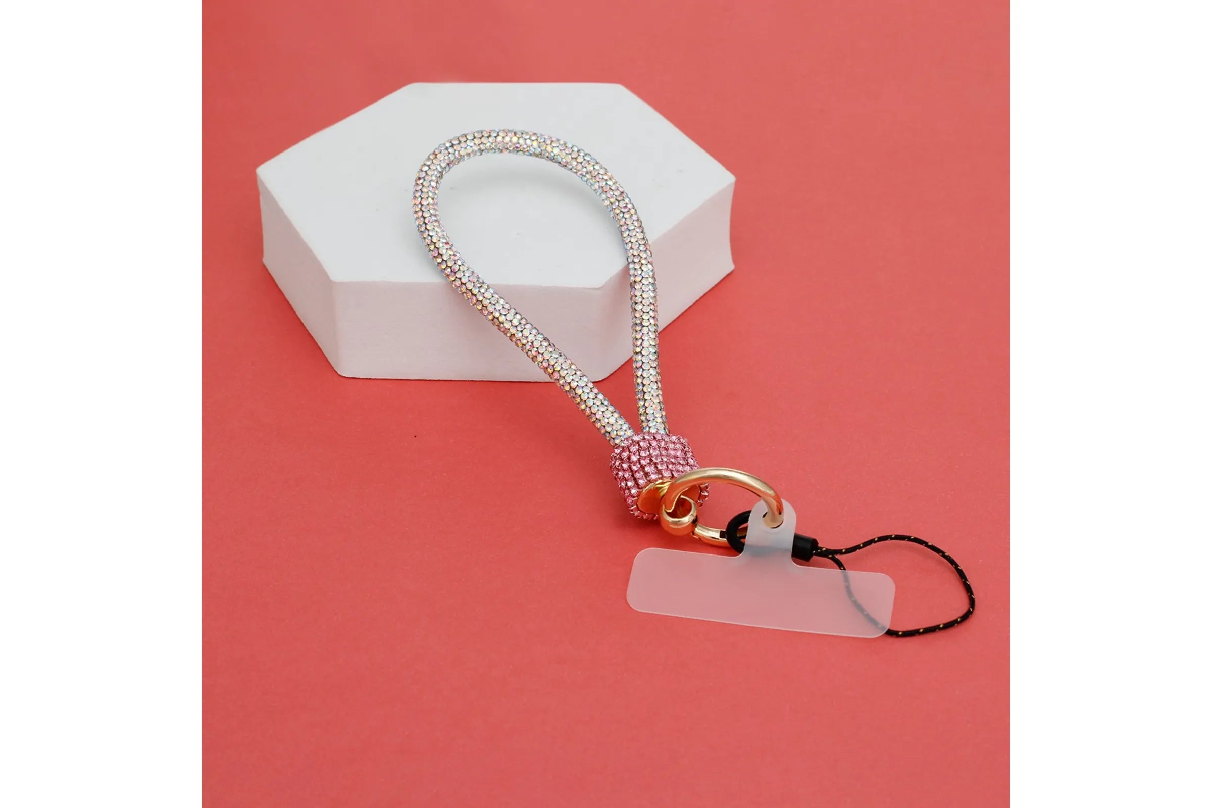 Pink and aurora borealis rhinestone-encrusted rope phone wrist strap draped over white hexagonal display prop on coral red background, showing full loop, pink rhinestone barrel wrap, round gold carabiner clasp, white plastic card insert, and black cord loop