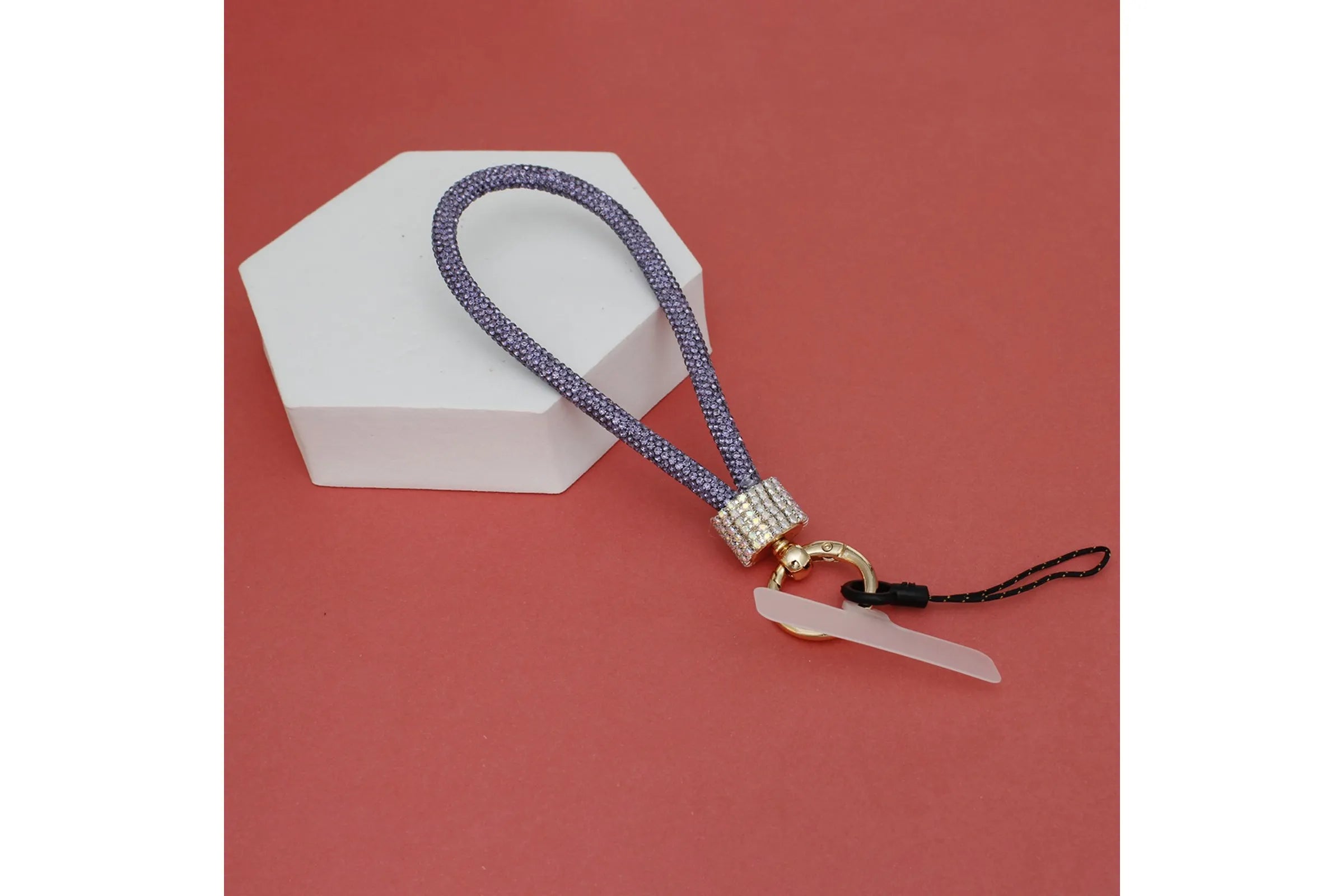 Purple rhinestone-encrusted rope phone wrist strap draped over white hexagonal display prop on red background, showing full loop, clear rhinestone barrel wrap, round gold carabiner clasp, clear plastic card insert, and black cord loop
