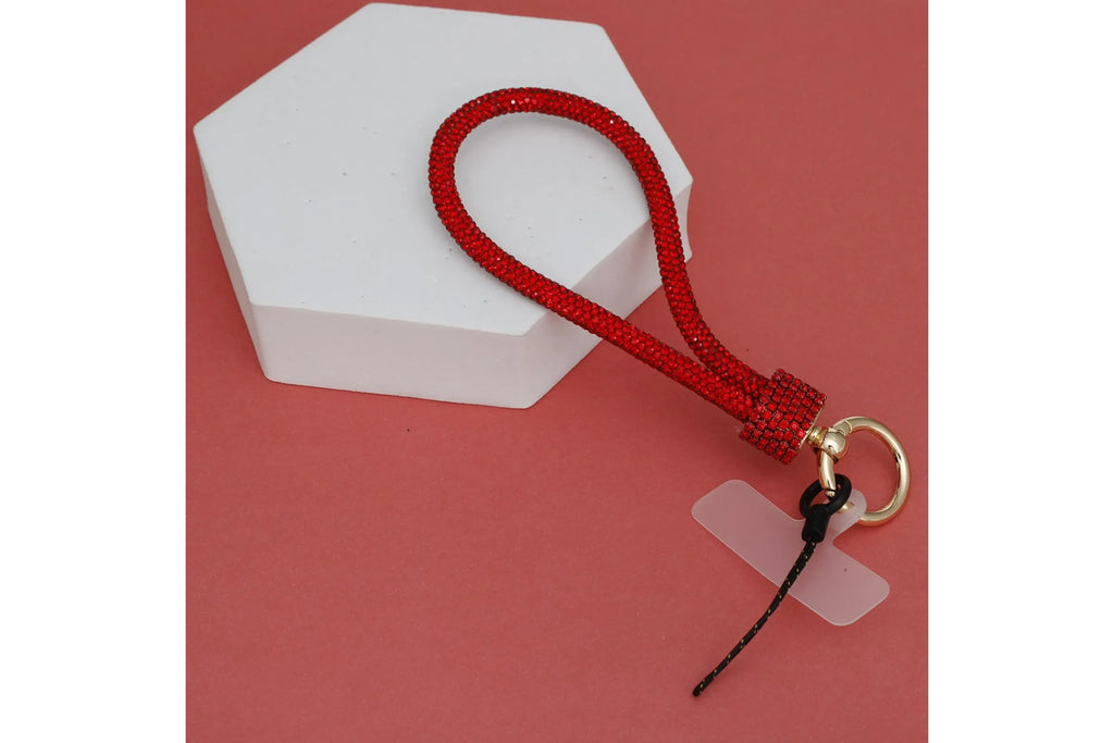 Red rhinestone-encrusted rope phone wrist strap draped over white hexagonal display prop on red background, showing full loop, red rhinestone barrel knot wrap, round gold carabiner clasp, clear plastic card insert, and black cord loop