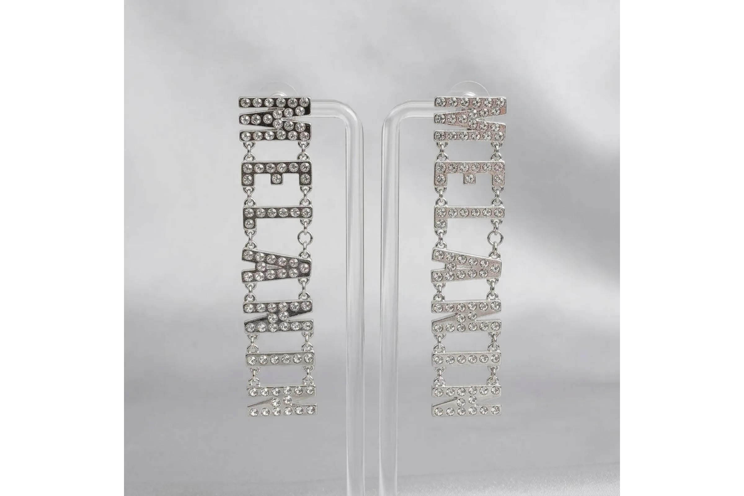 Pair of silver clear rhinestone MELANIN linked letter dangle earrings displayed on clear acrylic double T-bar earring stand against silver metallic background, showing full letter chain detail and post backings