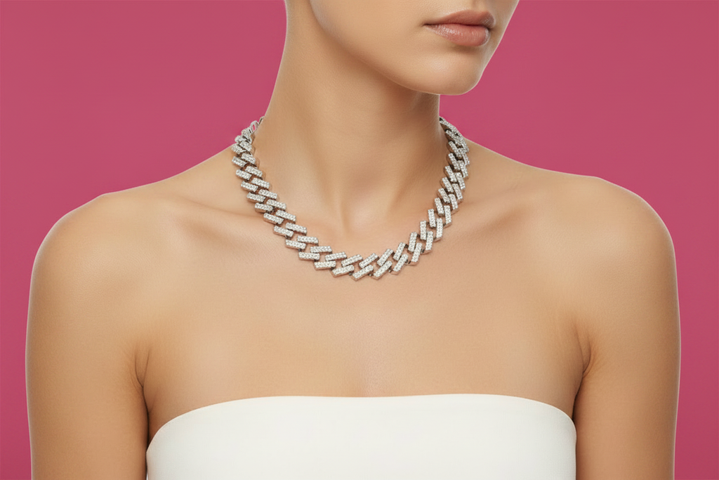 A woman with light skin wearing a white strapless top poses against a bold magenta-pink background. She wears a wide silver tone fully pave clear rhinestone square Cuban chain link necklace sitting at the collarbone, the interlocking square links sparkling under soft studio lighting.