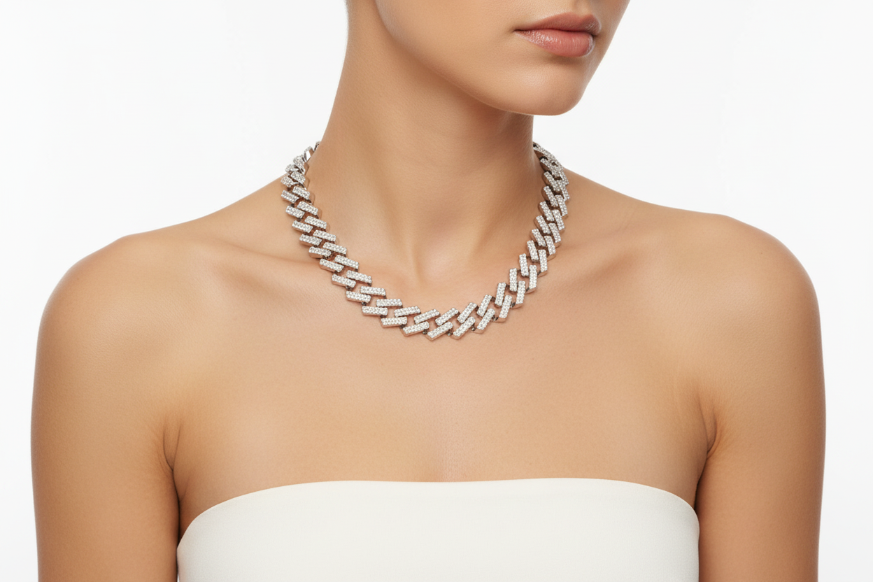 A woman with light skin wearing a white strapless top poses against a soft white background. She wears a wide silver tone fully pave clear rhinestone square Cuban chain link necklace sitting at the collarbone, the chain links clearly visible and sparkling.