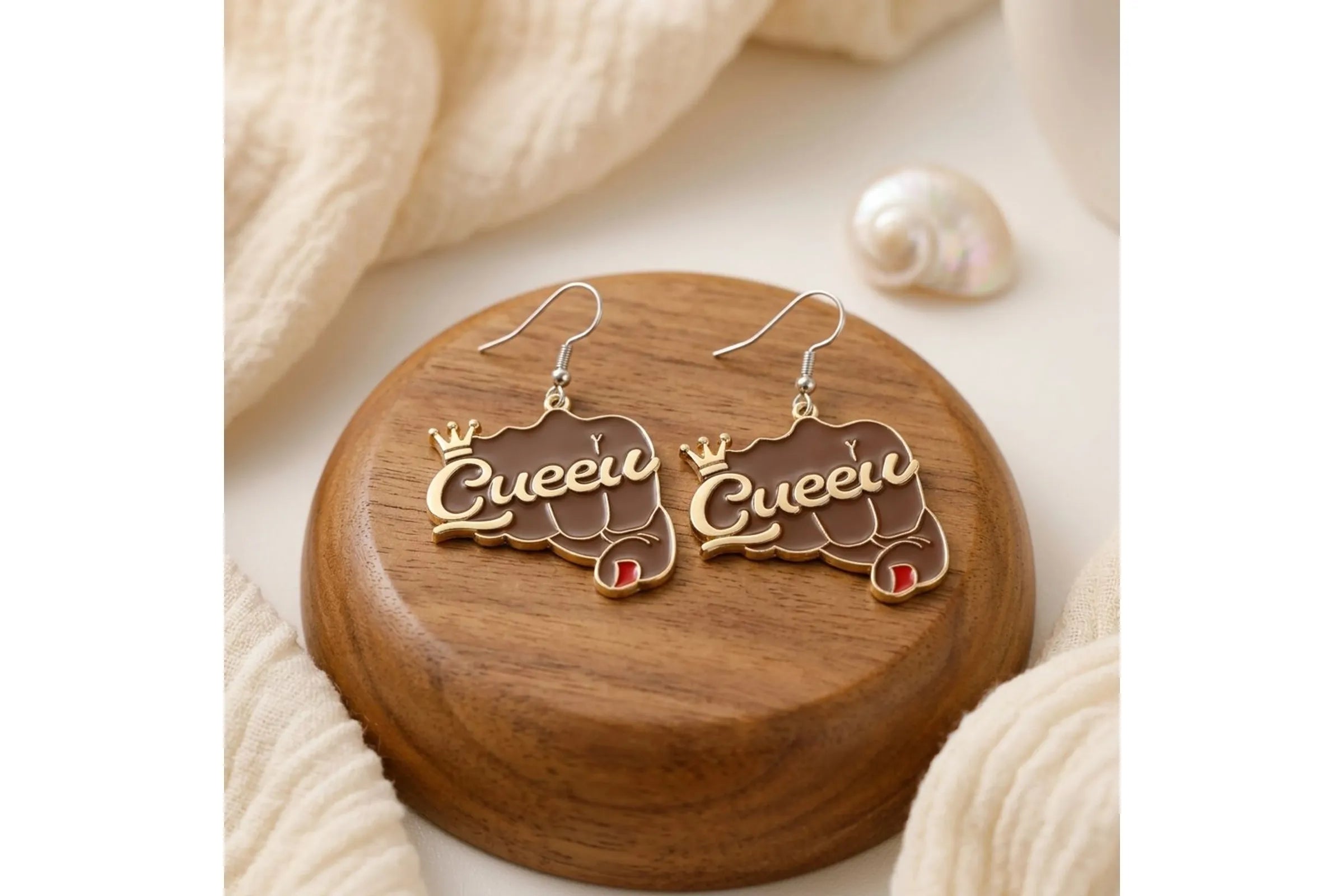 Pair of brown epoxy raised fist "Queen" crown fish hook dangle earrings displayed on round wooden jewelry prop with cream knit fabric and small shell in background