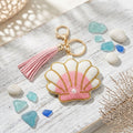 Pink and white seed bead seashell charm keychain with pearl accent, pink faux leather tassel, and gold lobster clasp, styled on whitewashed wood surface with sea glass and coral on white background
