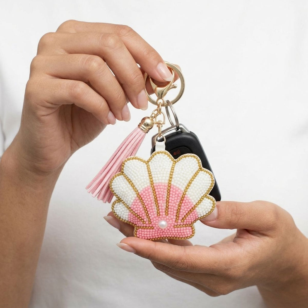 Pink and white seed bead seashell charm keychain with pearl accent, pink tassel, and gold lobster clasp, held in model's hands with a car key fob on white background