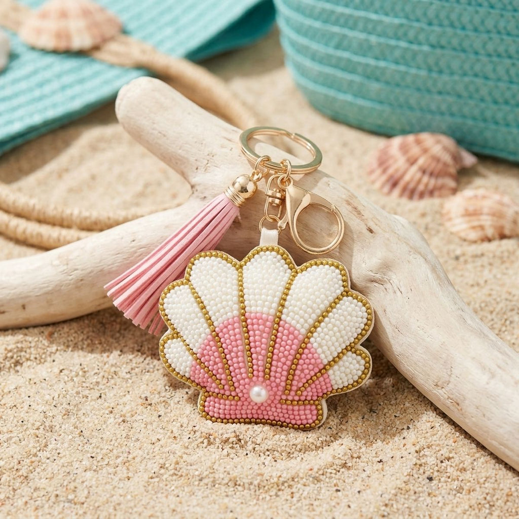 Pink and white seed bead seashell charm keychain with pearl accent, pink faux leather tassel, and gold lobster clasp, resting on sand with driftwood and natural seashells