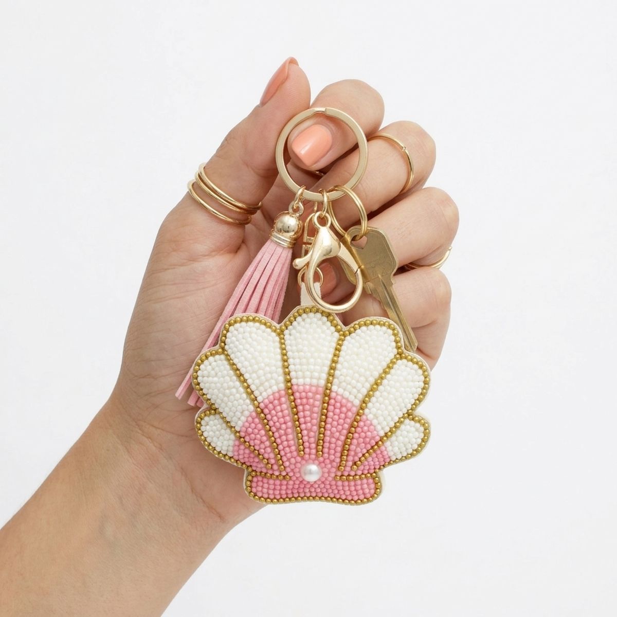 Pink and white seed bead seashell charm keychain with pearl accent, pink faux leather tassel, and gold lobster clasp, held in model's hand with gold rings and a house key on white background