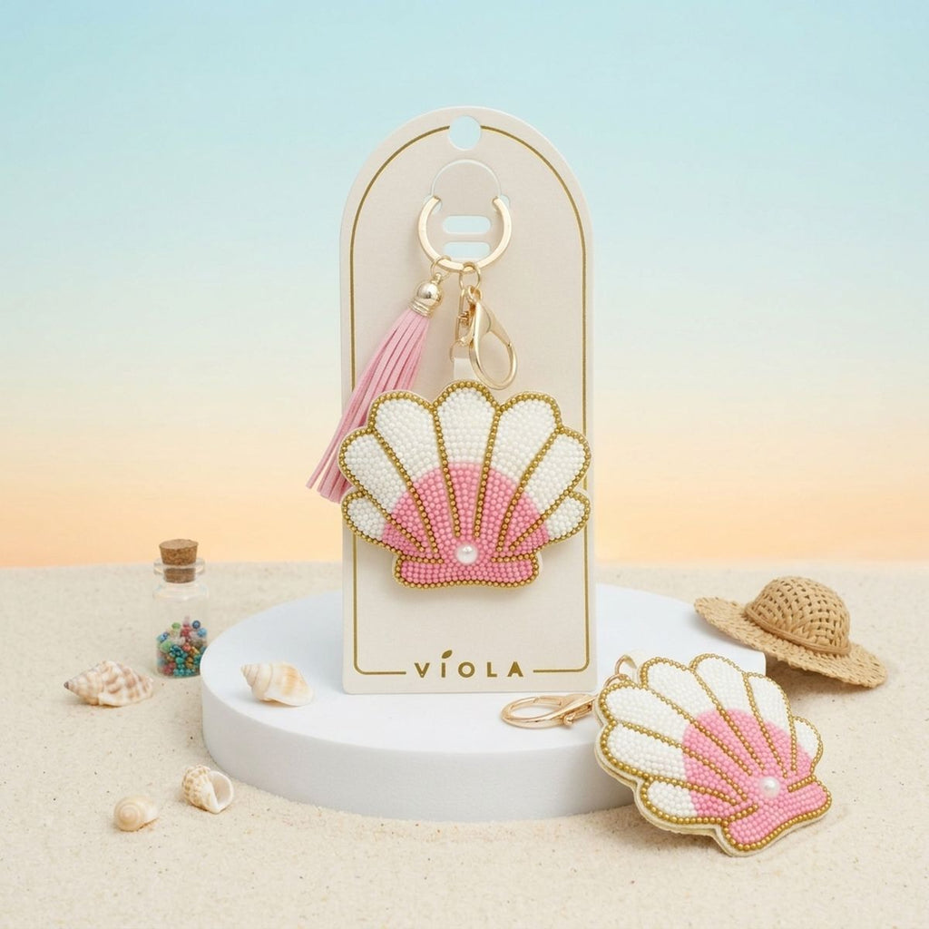 Pink and white seed bead seashell charm keychain with pink tassel displayed on packaging card, shown front and back alongside miniature straw hat and seashell props on sand with sunset gradient background
