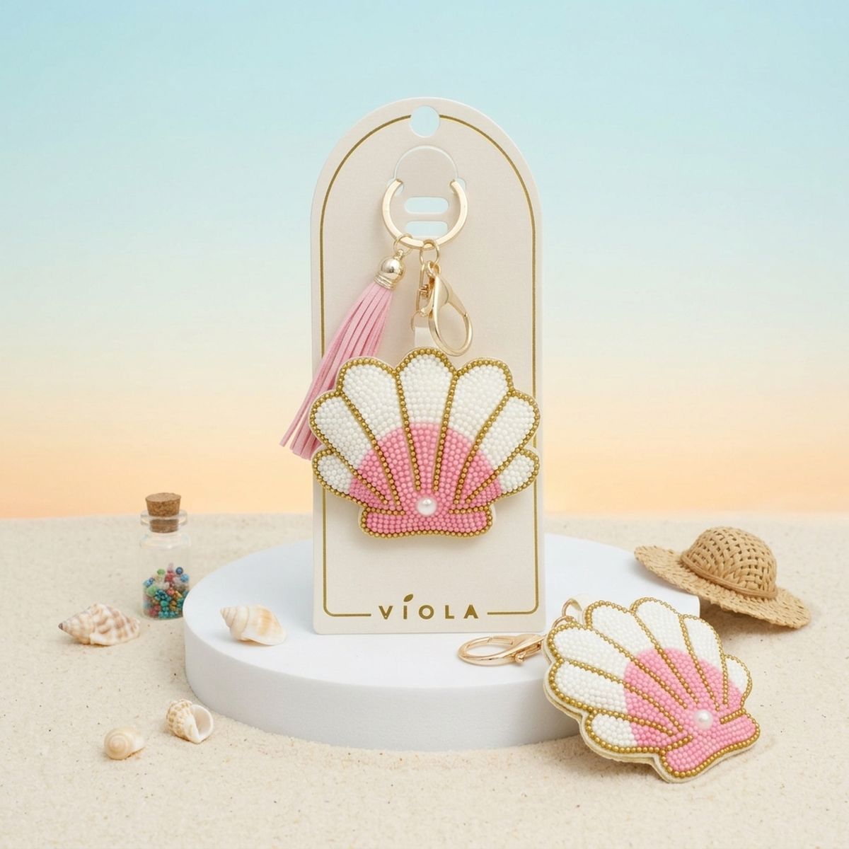 Pink and white seed bead seashell charm keychain with pink tassel displayed on packaging card, shown front and back alongside miniature straw hat and seashell props on sand with sunset gradient background