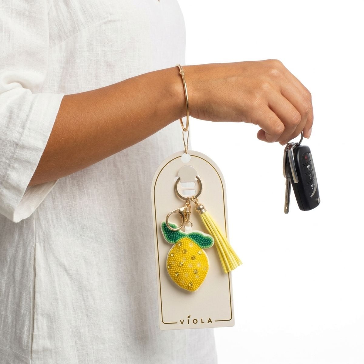 Yellow and green seed bead lemon charm keychain with yellow faux leather tassel and gold lobster clasp, shown on packaging card held by model in white linen top