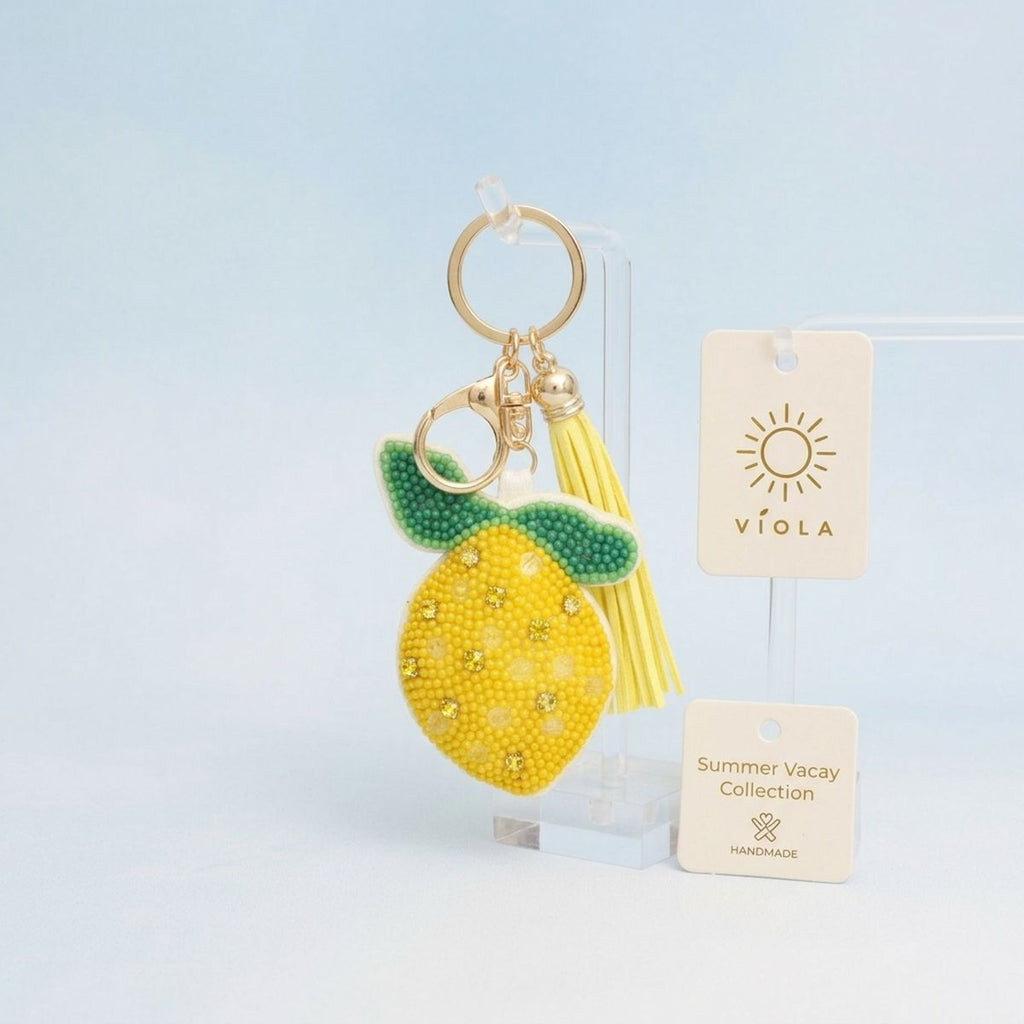 Yellow and green seed bead lemon charm keychain with rhinestone accents, yellow faux leather tassel, and gold lobster clasp, displayed on acrylic stand with light blue background