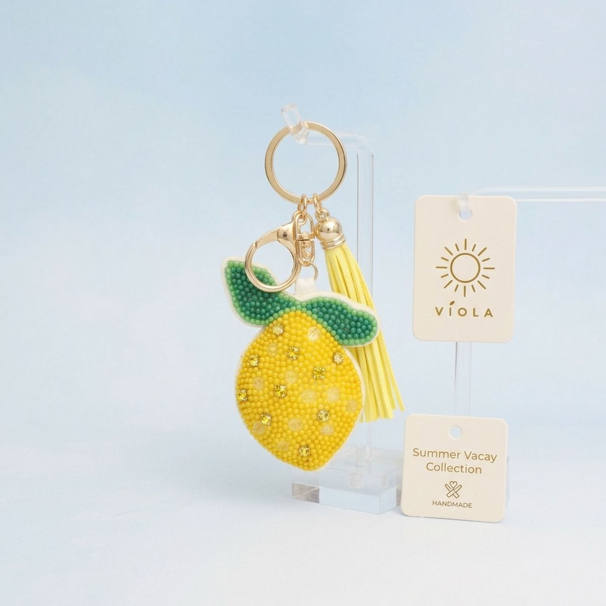 Yellow and green seed bead lemon charm keychain with rhinestone accents, yellow faux leather tassel, and gold lobster clasp, displayed on acrylic stand with light blue background