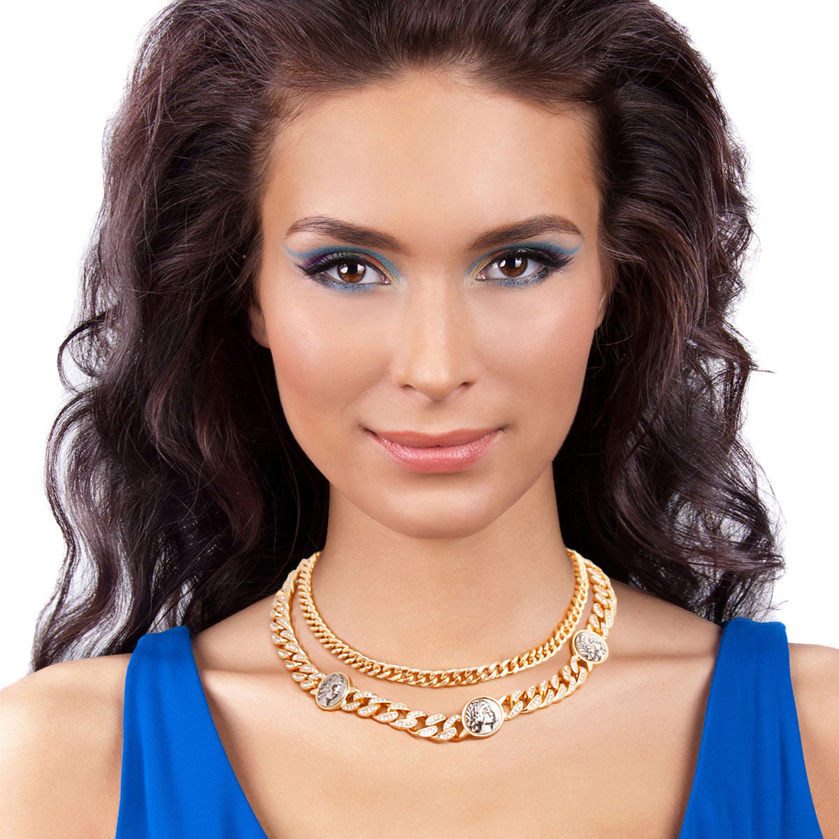 A woman with medium skin, long dark wavy hair, and bold blue eye makeup poses against a white background wearing a cobalt blue sleeveless top. She wears a 2-layer rhinestone pave Cuban chain necklace in gold tone at the collarbone, with three burnished antique gold coin medallions featuring a Roman profile face detail spaced along the lower chain.